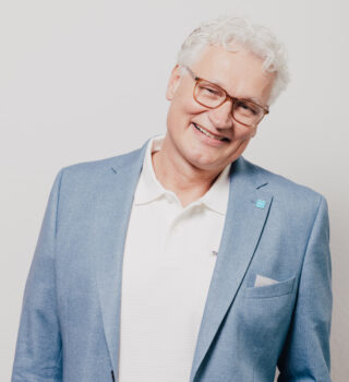 Ein Mann namens Claas Ohnesorge mit grauen kurzen Haaren und eine Brille. Er trägt einen hellblauen Anzug und einen weißen Pullover.