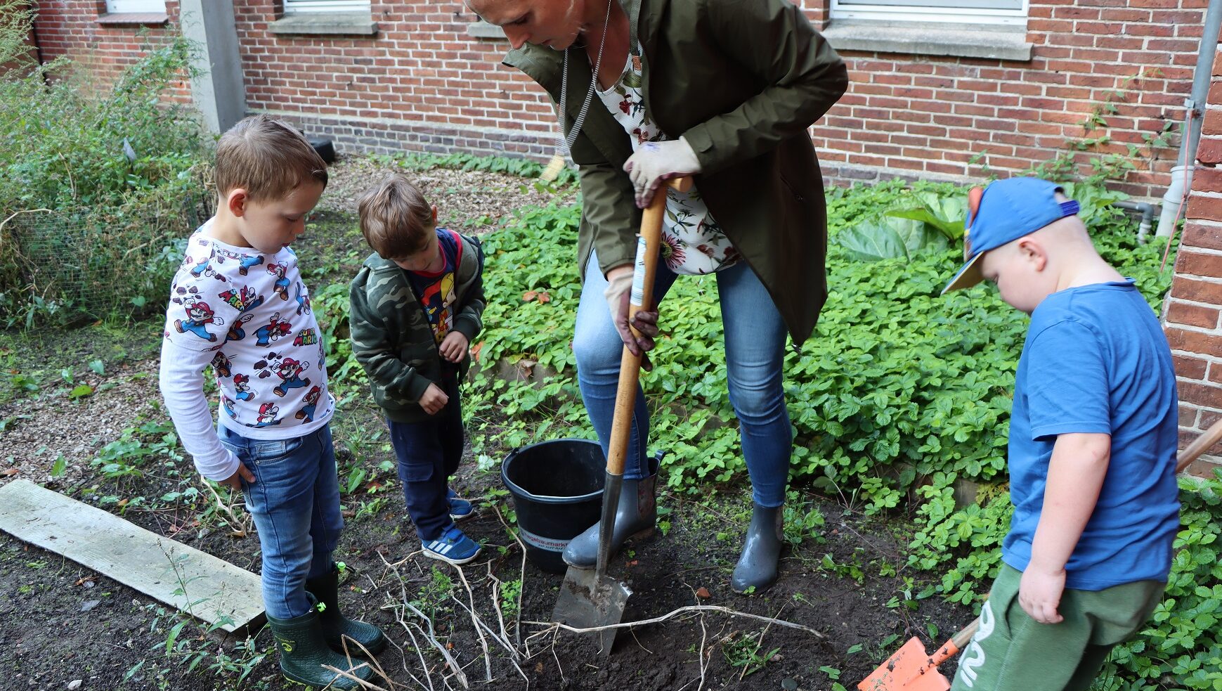 Drei kleine Kinder und eine Frau sind draußen im Garten. Die Frau mach ein Loch mit einer Schaufel auf dem Beet.