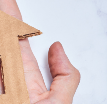 Eine Hand mit ein gebasteltes Häuschen aus brauner Pappe. Man sieht auch einen weißen Hintergrund.