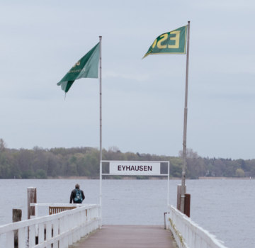 Draußen am Meer. Man sieht im Hintergrund einen Wald und einen langen Steg am Wasser. Auch sieht man zwei Flaggen und ein Schild mit der Aufschrift "Eyhausen"