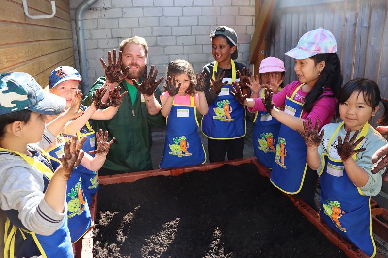 Ein Mann (Gärtner) mit acht kleinen Kindern stehen vor einem Beet mit Sand. Sie halten alle ihre schmutzigen Hände hoch.