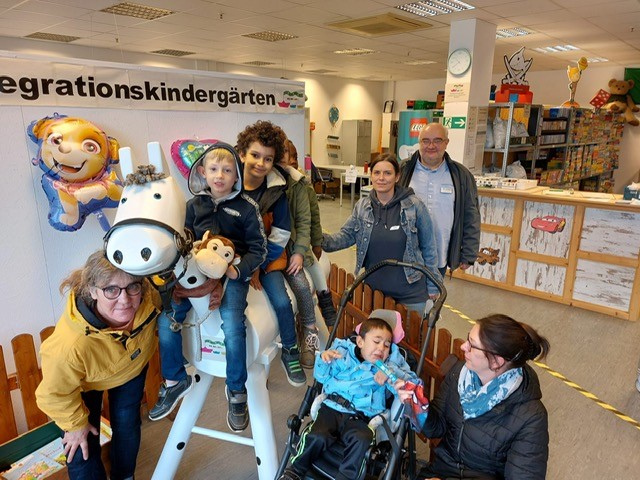 Drei Frauen, ein Mann und vier Kinder sind auf dem Bild zu sehen. Man sieht auch eine Wand mit der Aufschrift "Integrationskindergärten".