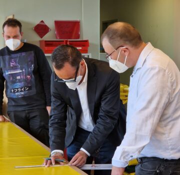 Zwei GPS Beschäftigte und ein Politiker. Der Politiker misst etwas aus mit einem Zollmaßstab. Der Politiker soll die Arbeit der Beschäftigten erleben.