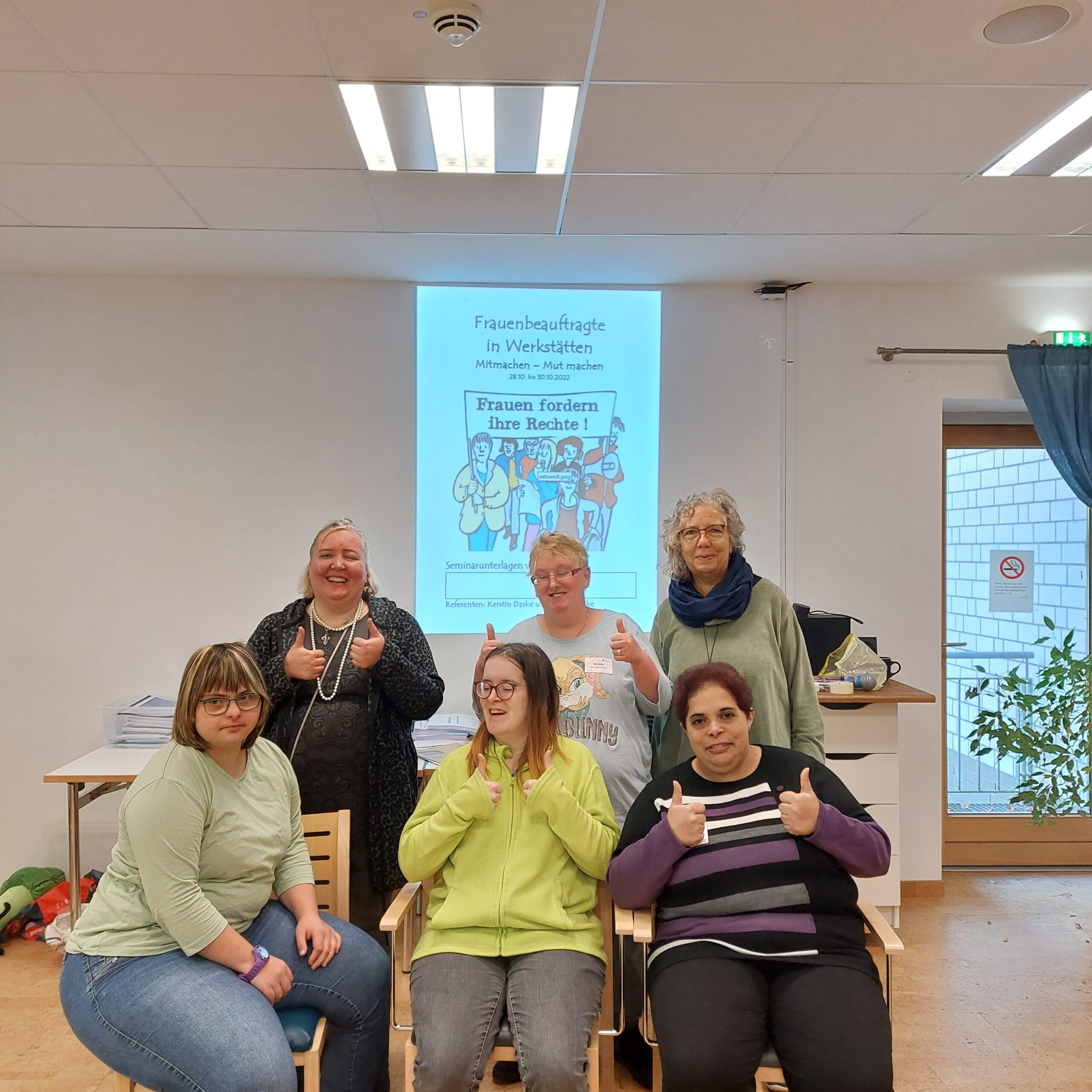 Gruppenfoto Frauenbeauftragte Andrea Hicken und fünf GPS Beschäftigte sind in einem Raum. Sie stehen oder sitzen. Im Hintergrund an der Leinwand sie man ein Bild mit dem Thema "Frauenbeauftragte in Werkstätten - Frauen fördern ihre Rechte!" Sie halten alle ihre Daumen hoch.