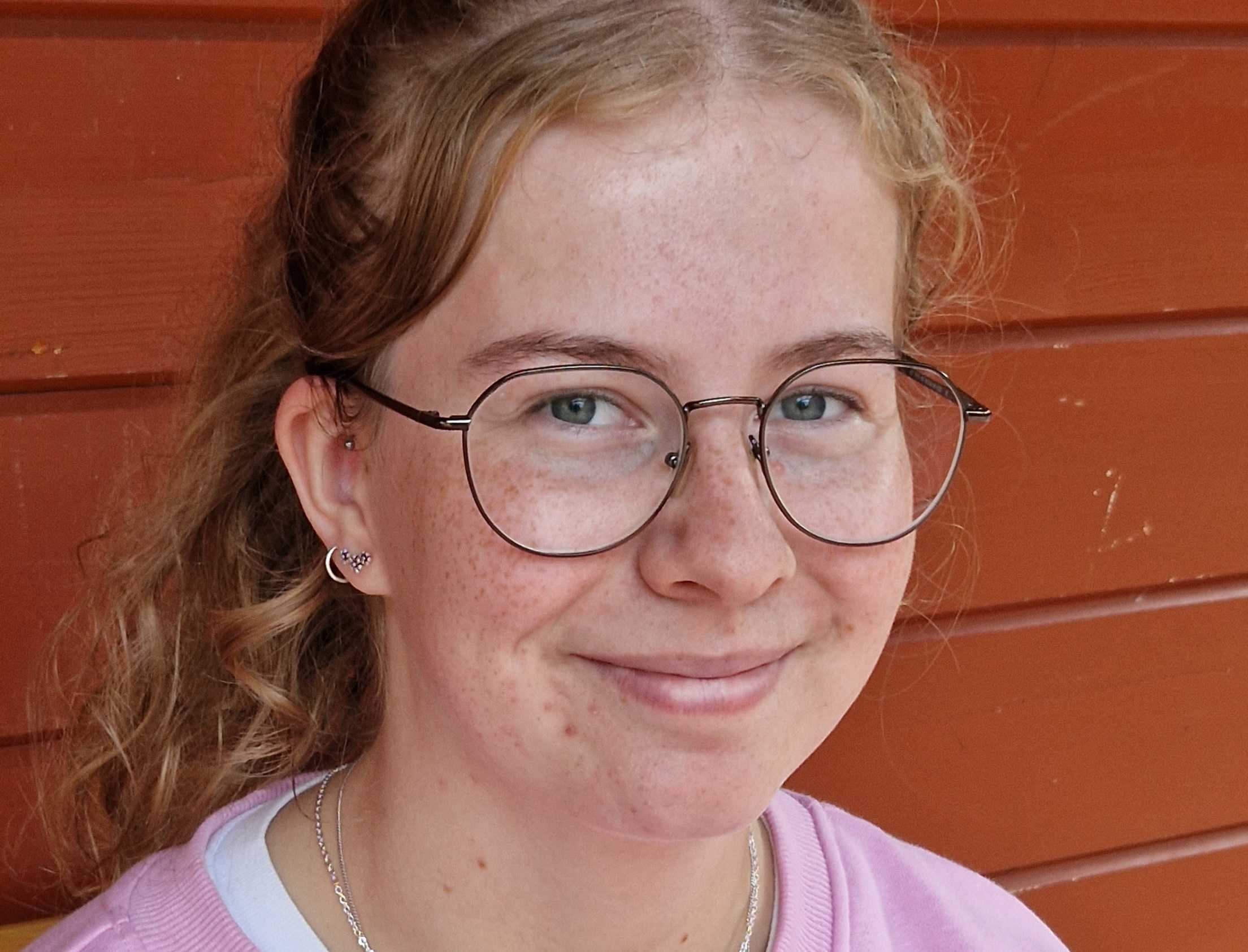 Eine junge Frau lächelt in die Kamera. Sie steht an einer roten Wand.