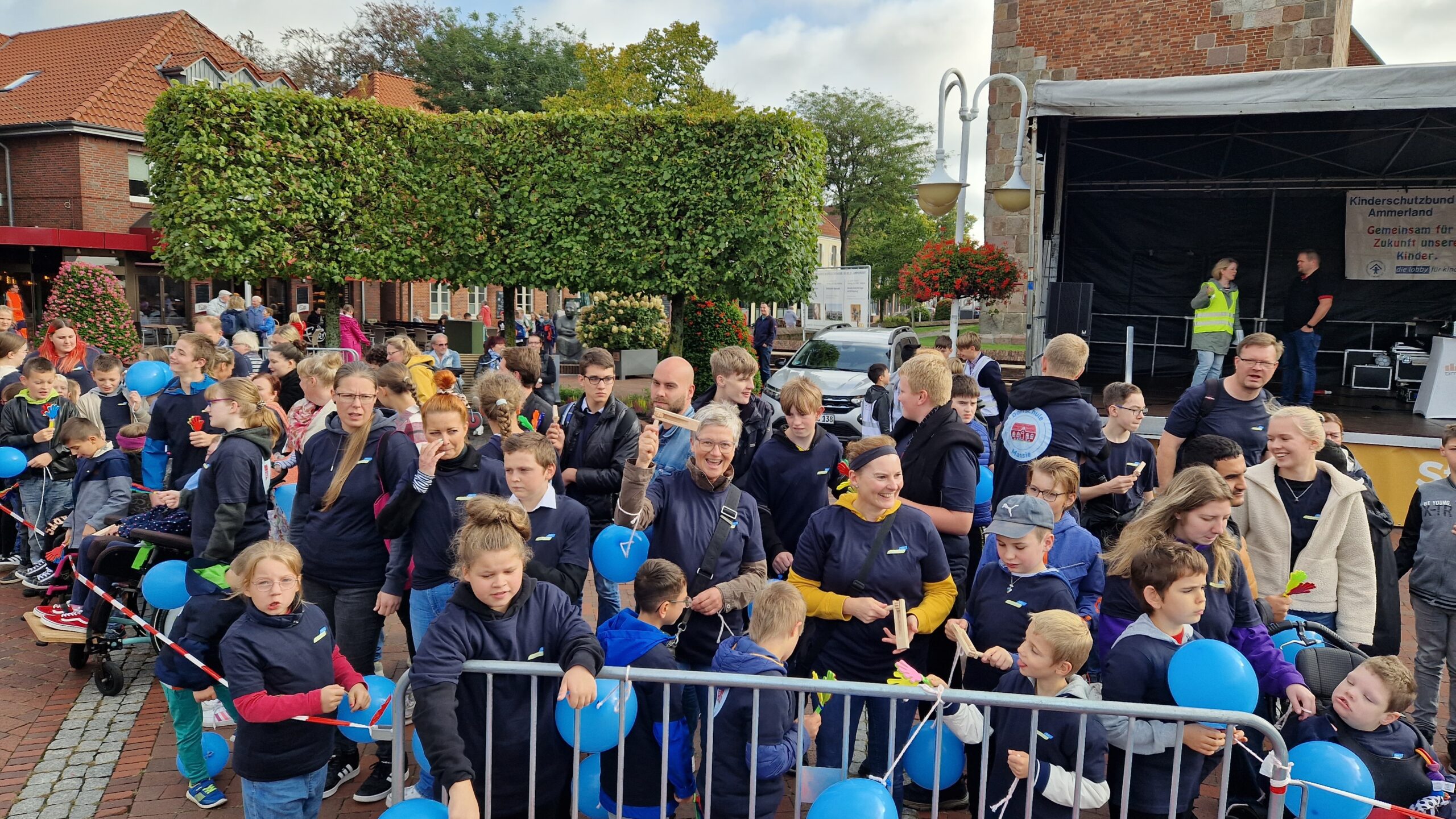 Man sieht draußen Erwachsene, Kinder und Jugendliche bei einem Fest. Im Hintergrund sieht man auch eine Bühne.