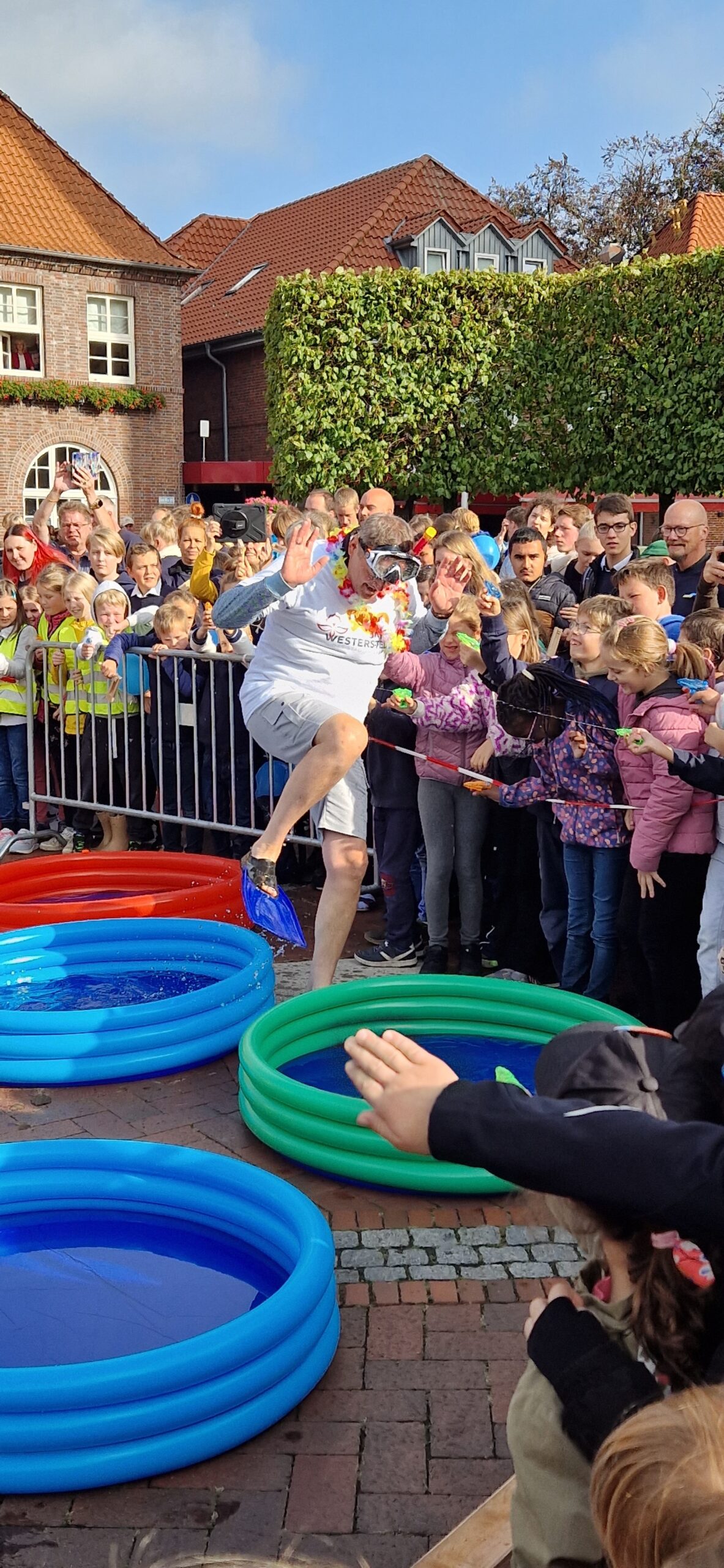 Auf diesem Bild sieht man Kinder draußen im Hintergrund. Ein Mann muss eine Hüpfwette in kleine Swimmingpools machen.