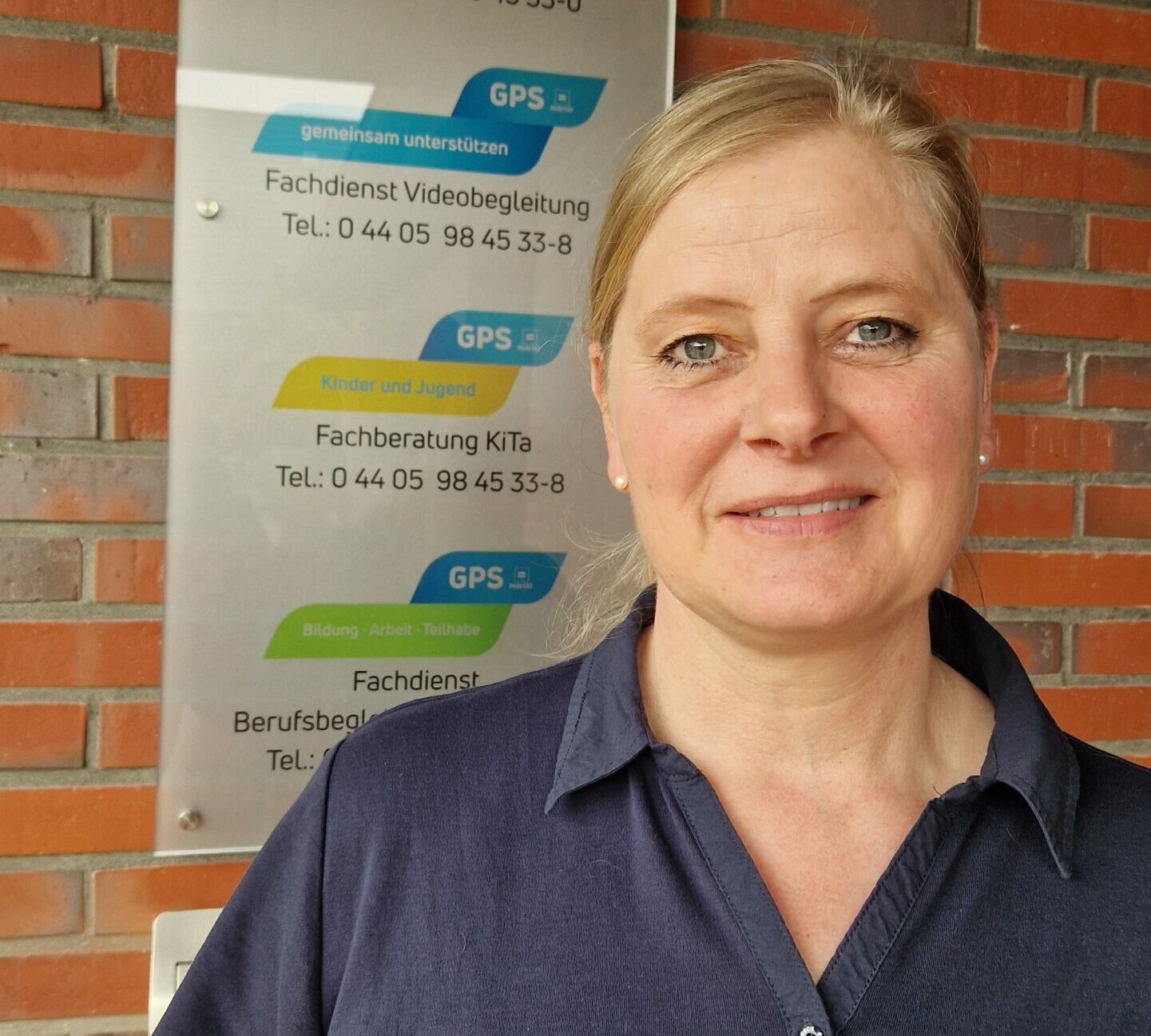 Eine GPS Mitarbeiterin schaut in die Kamera vor einem Eingang eines Gebäudes der GPS Beratungsstellen. Man sieht im Hintergrund eine Infotafel, wo Beratungsstellen von drei Geschäftsbereichen der GPS darauf abgebildet sind.