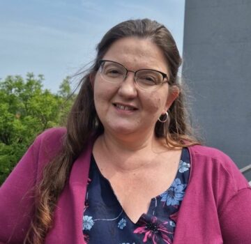 Eine junge Frau steht draußen vor der Kamera. Sie schaut freundlich.