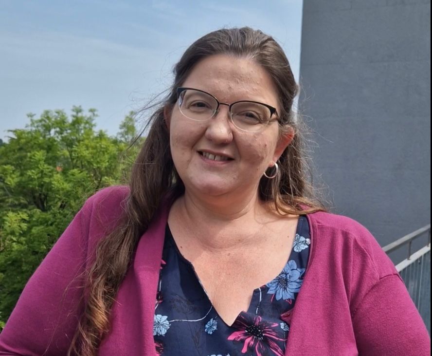 Eine junge Frau steht draußen vor der Kamera. Sie schaut freundlich.