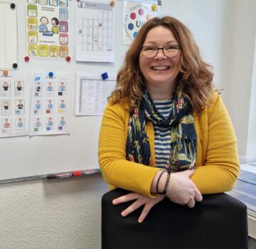 Eine GPS Mitarbeitern names Sandra Yvonne Dreyer von der Abteilung "Unterstütze Kommunikation" in einem Raum. Sie lächelt in die Kamera. Im Hintergrund sieht man eine Flip-Chart mit vielen angehängten Dingen.