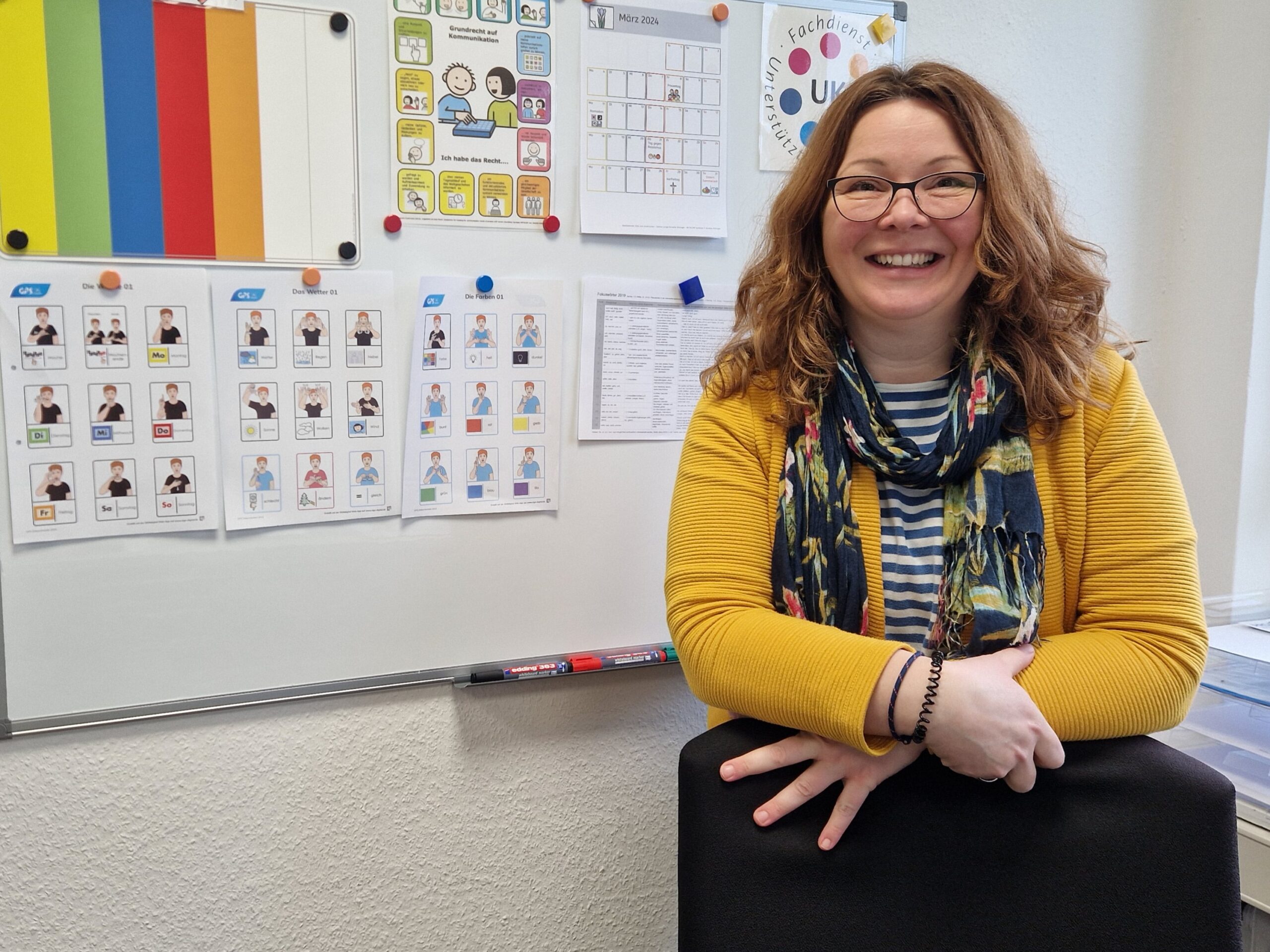 Eine GPS Mitarbeitern names Sandra Yvonne Dreyer von der Abteilung "Unterstütze Kommunikation" in einem Raum. Sie lächelt in die Kamera. Im Hintergrund sieht man eine Flip-Chart mit vielen angehängten Dingen.