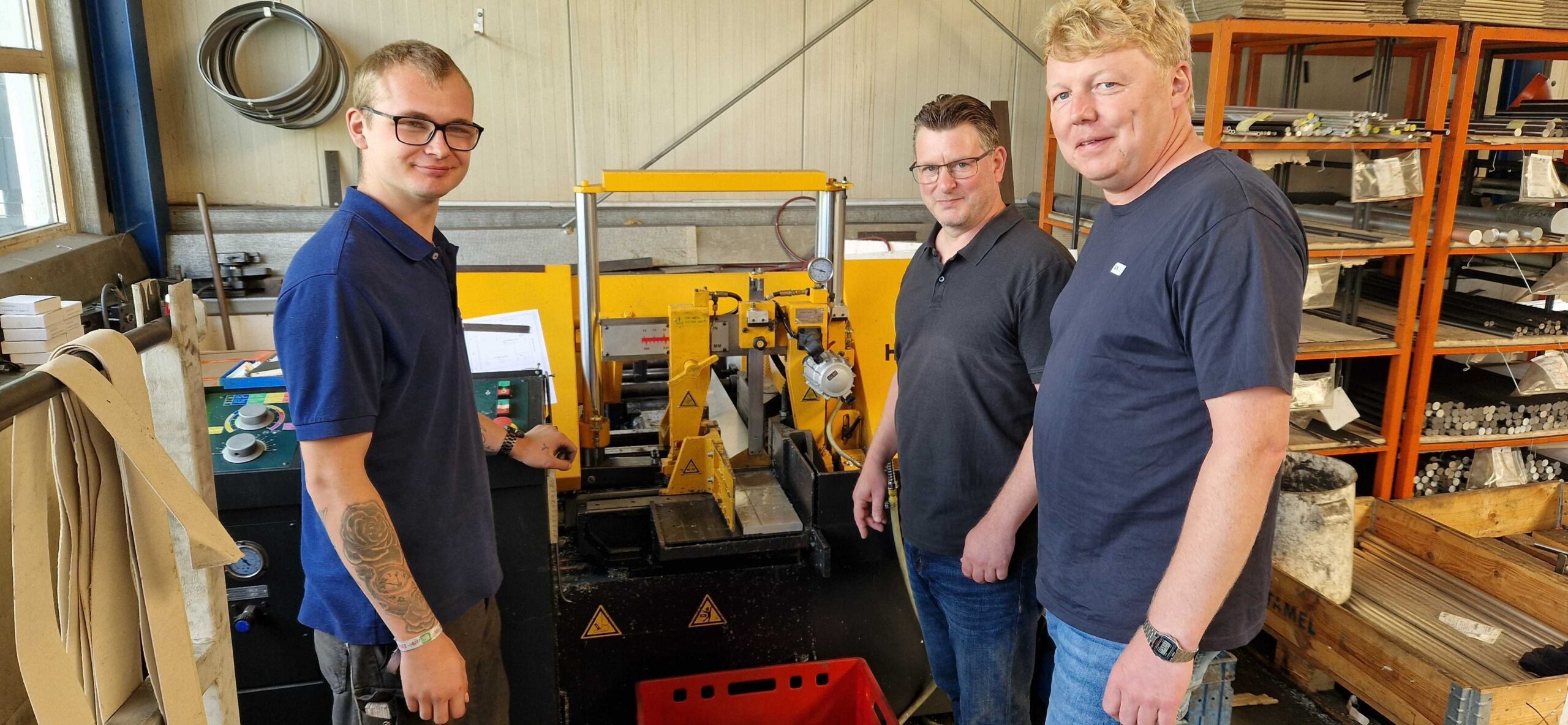 Gruppenfoto mit zwei Männern und einen jungen Mann vor einer Maschine in einer Werkstatt.