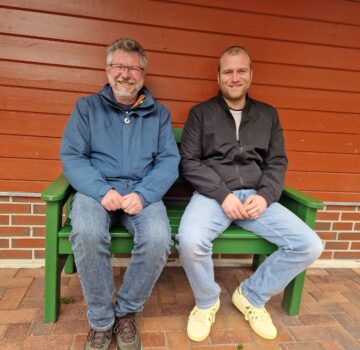 Zwei GPS Mitarbeiter Hans-Hermann Kickler und Thore Opitz sitzen auf einer Bank vor der Wand eines Hauses. Sie lächeln freundlich in die Kamera.