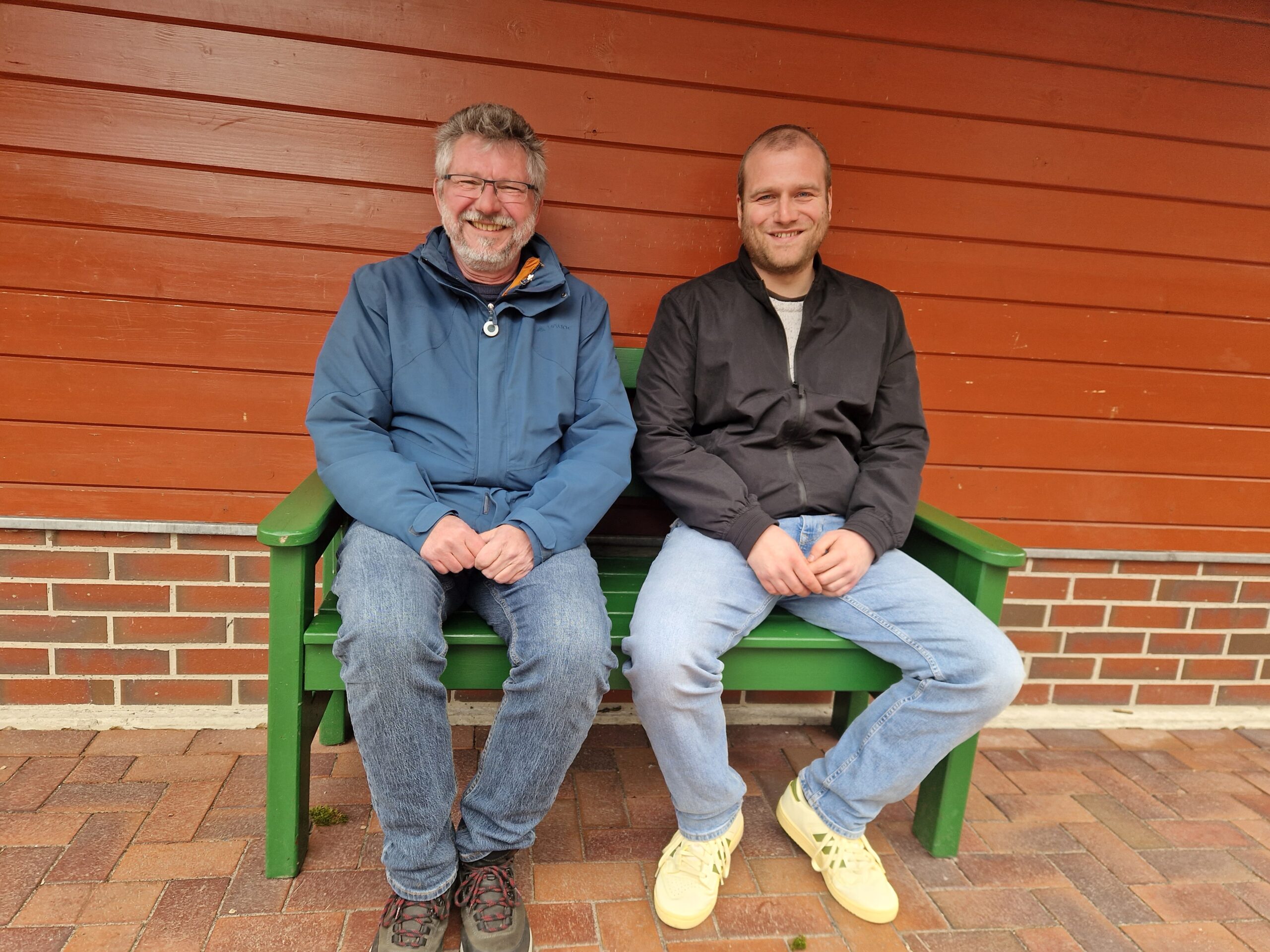 Zwei GPS Mitarbeiter Hans-Hermann Kickler und Thore Opitz sitzen auf einer Bank vor der Wand eines Hauses. Sie lächeln freundlich in die Kamera.