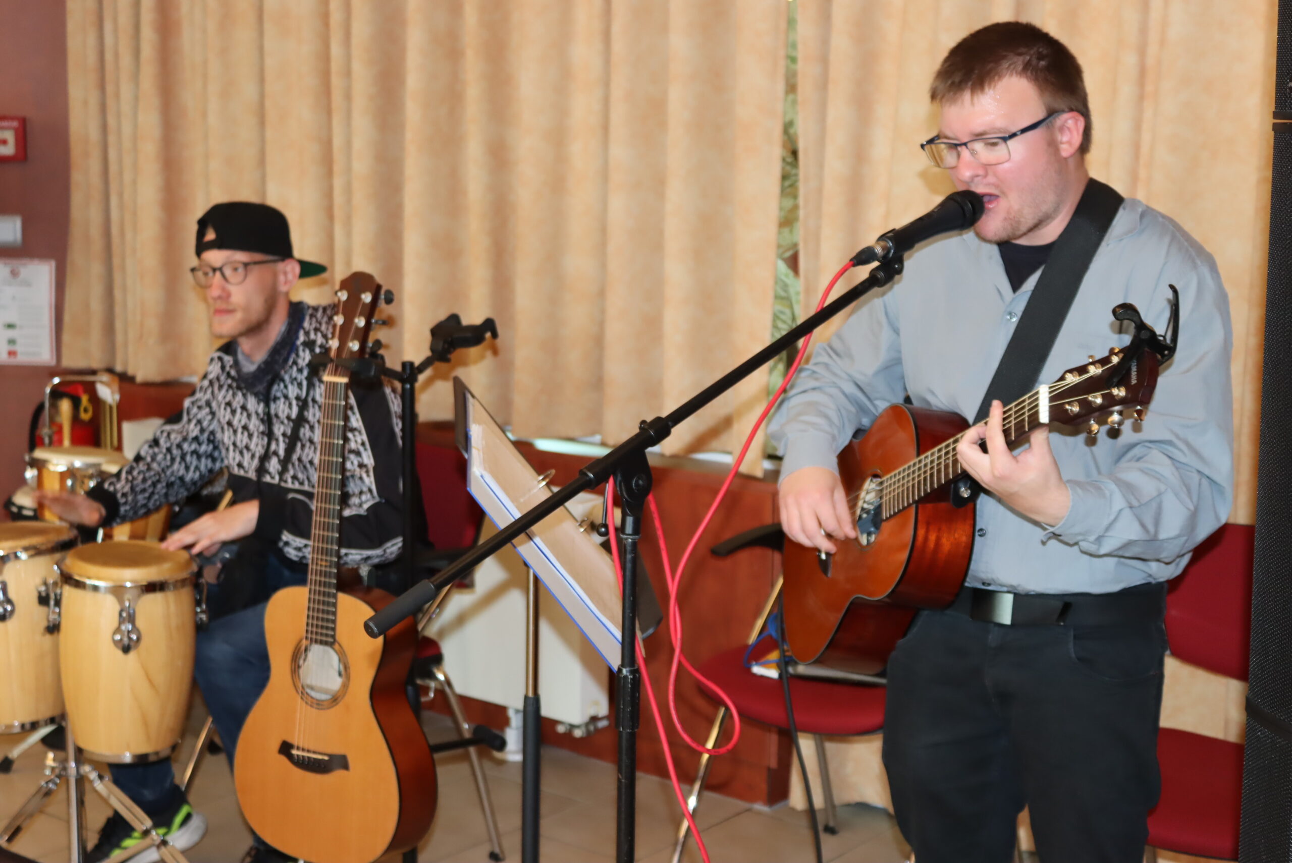 Ein junger Mann spielt Gitarre und singt an einem Mikrofon. Ein anderer junger Mann sitzt und spielt mit ein anderes Musikinstrument.