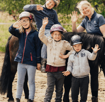 Eine Gruppe von jungen Kindern mit einer Trainerin beim Pferde reiten.
