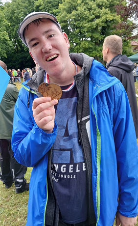 Ein junger Mann zeigt eine Medaille vor der Kamera. Viele Menschen sind im Hintergrund zu sehen.