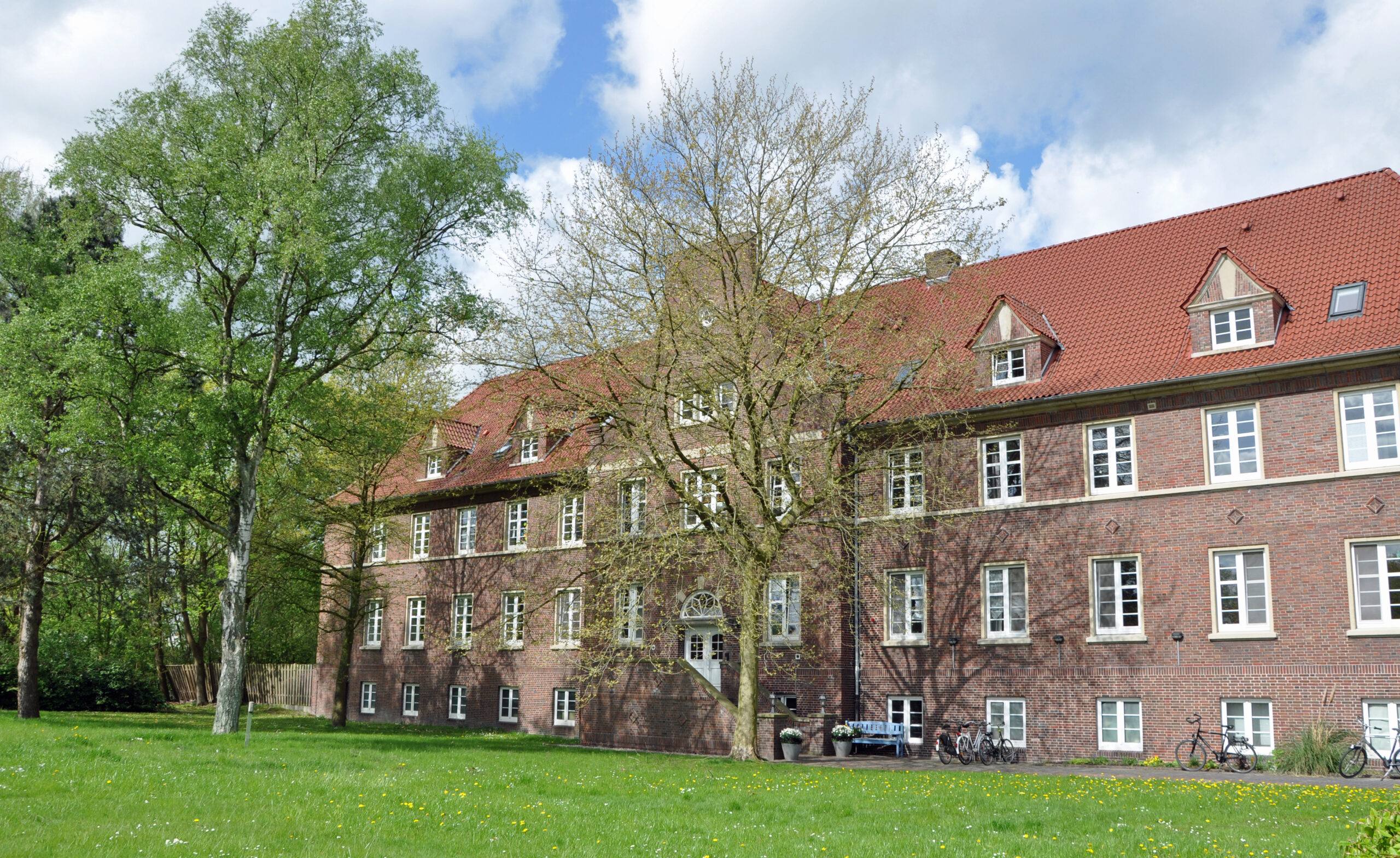 Ein "GPS Windrad 1-4 Gebäude" draußen im Vorgarten. Man sieht zum Beispiel Bäume, Fahrräder und einen großen Rasen. Draußen ist schönes Wetter.