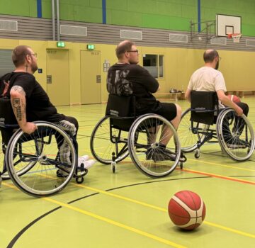 Fünf Menschen spielen Rollstuhl-Basketball in einer großen Sporthalle.