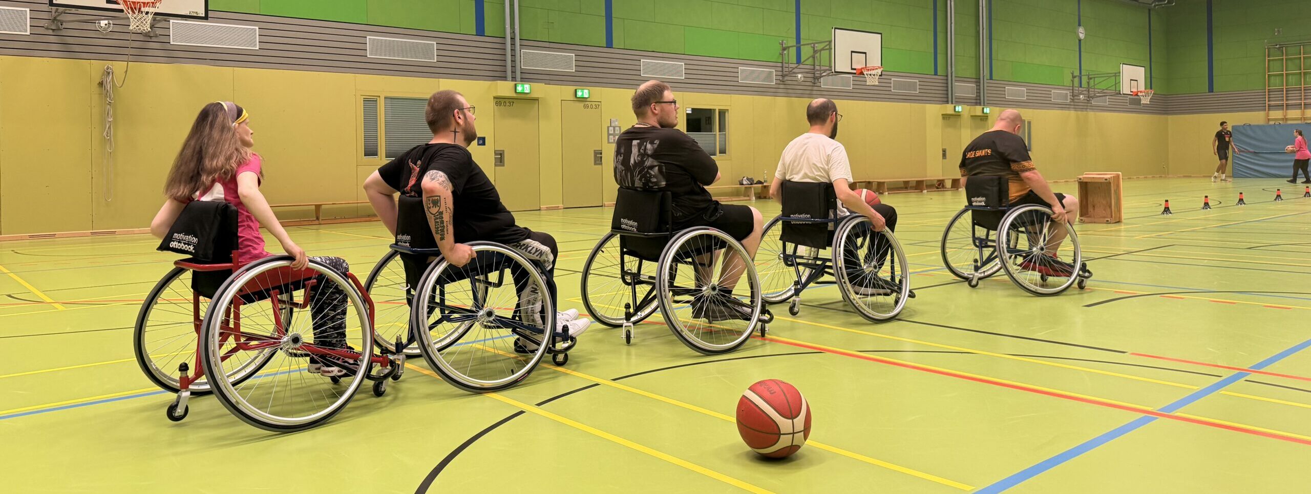 Fünf Menschen spielen Rollstuhl-Basketball in einer großen Sporthalle.