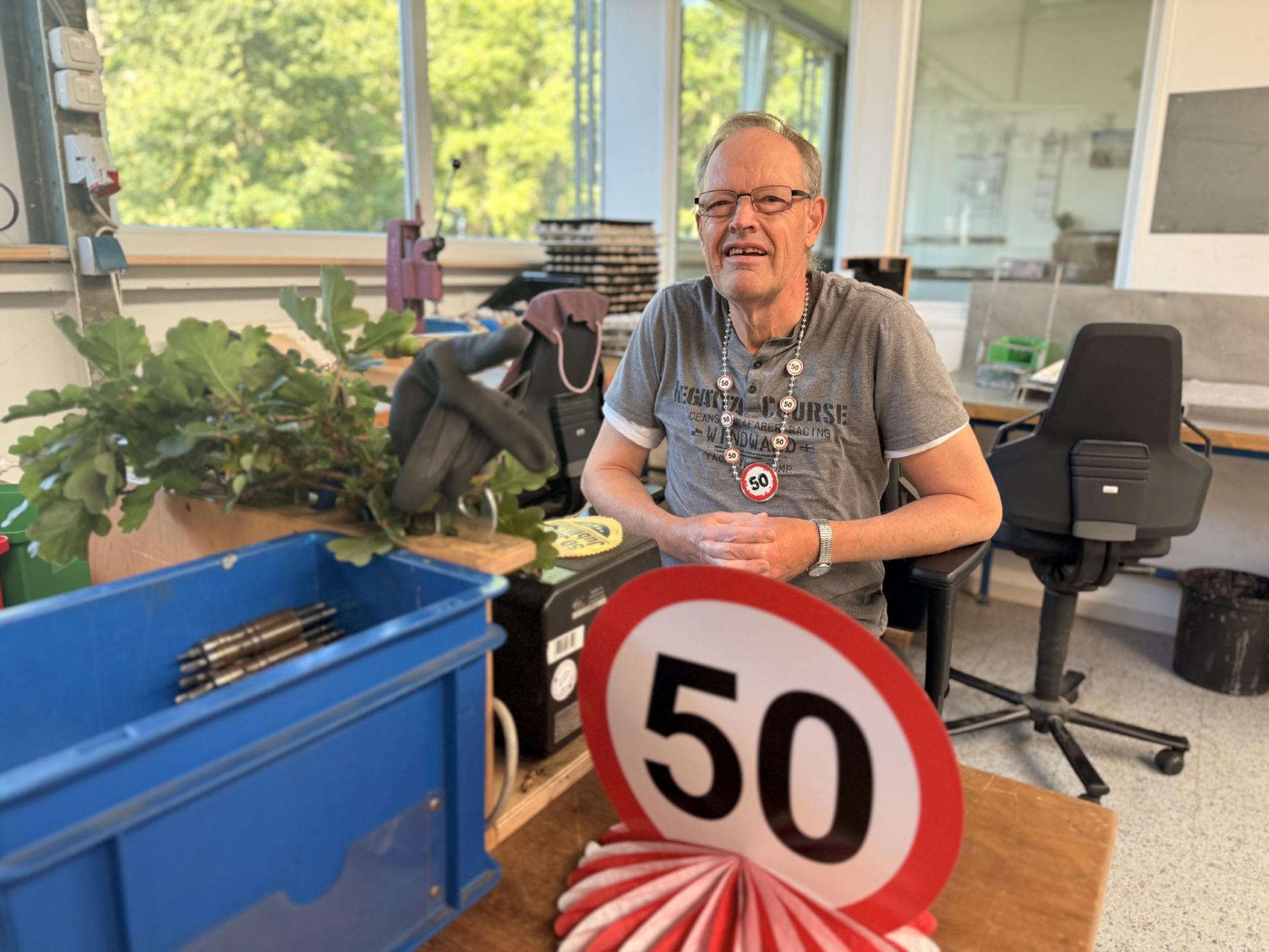 Ein GPS Beschäftigter feiert in der Werkstatt Jeringhave sein 50-jähriges Jubiläum. Er sitzt an seinem Arbeitsplatz. Man sieht ein rot-weißes Schild mit der Zahl "50".