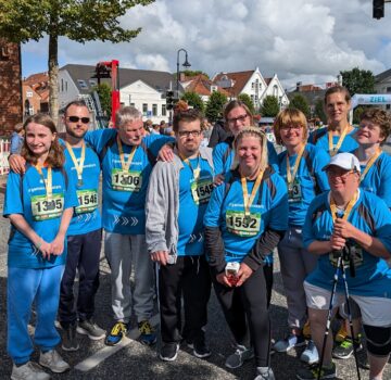Gruppenfoto draußen beim Sonnenschein. Eine Gruppe junger und ältere Menschen. Man sieht Zuschauer. Die Gruppe hat Sportsachen an.