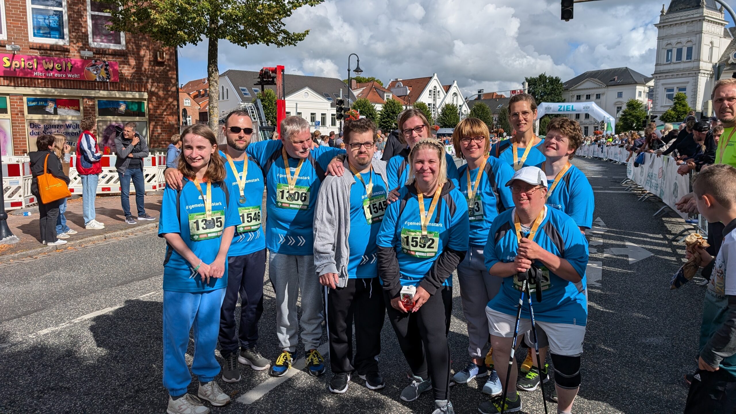 Gruppenfoto draußen beim Sonnenschein. Eine Gruppe junger und ältere Menschen. Man sieht Zuschauer. Die Gruppe hat Sportsachen an.