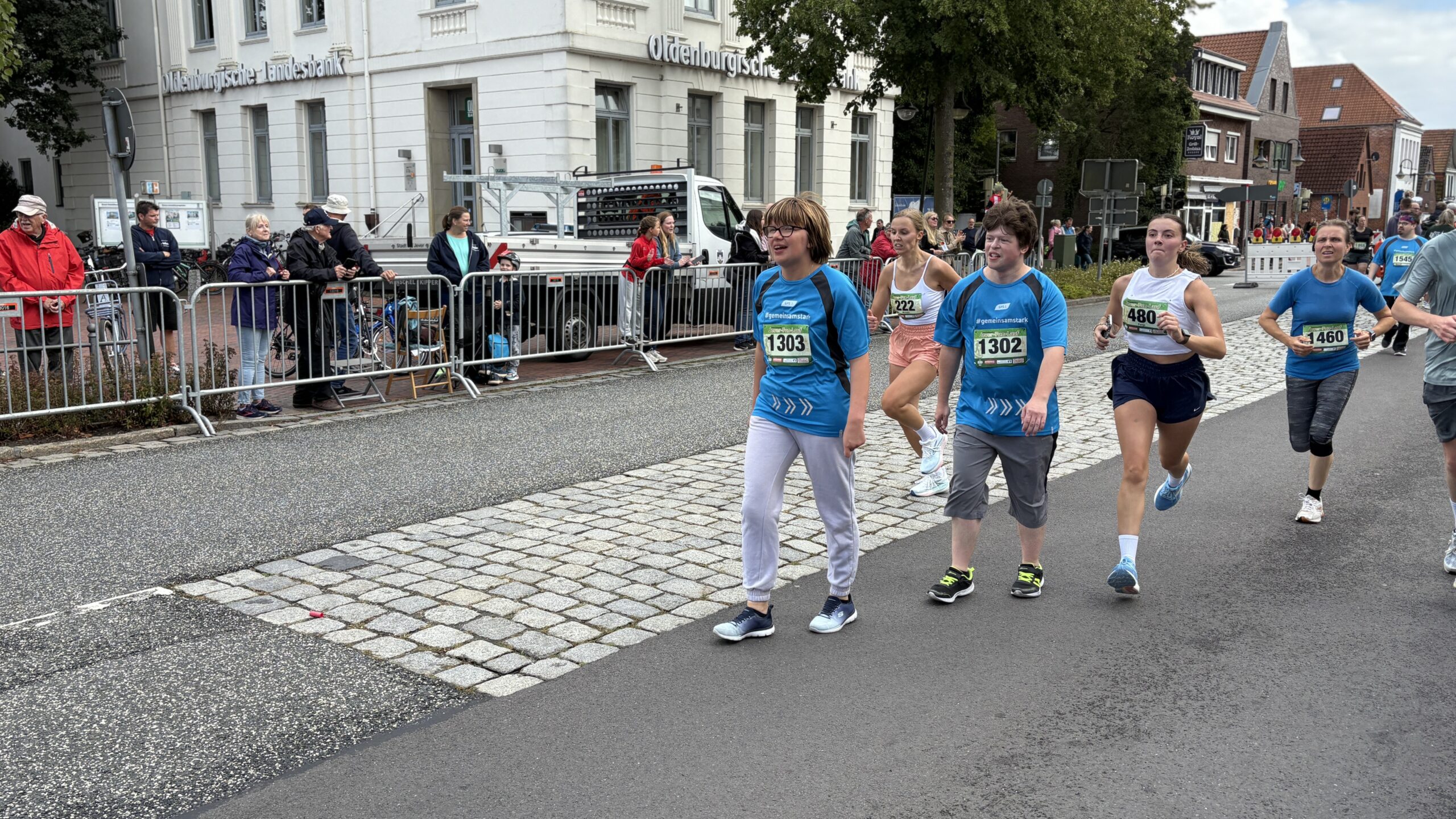 Junge Frauen und junge Männer machen ein Wettrennen draußen auf einer Straße. Zuschauer sehen ihnen zu.
