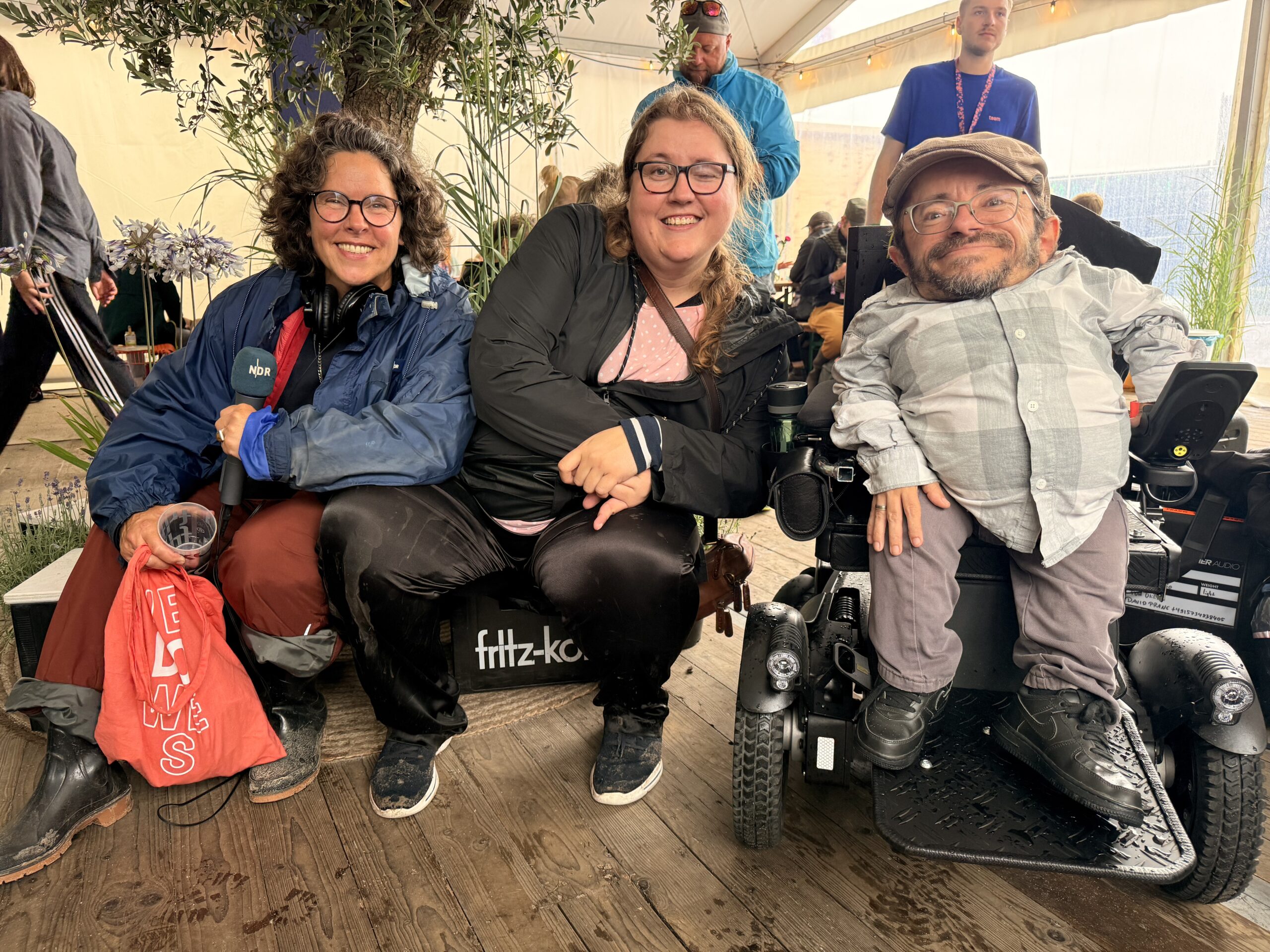 Anna-Helen Wippich vom Podcast „Kabelsalat“ sitzt gemeinsam mit Moderatorin Carola Schede (links) und dem Aktivisten Raúl Krauthausen (rechts) auf dem Watten Schlick Festival 2025 unter einem Pavillon. Alle drei lächeln in die Kamera. Im Hintergrund sieht man Menschen, Technik und das Zeltambiente des Festivals.