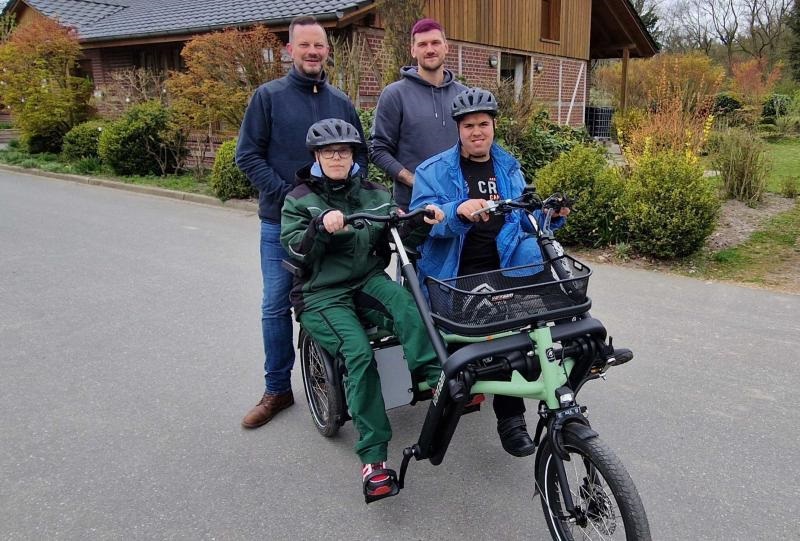 Ein Gruppenfoto mit einem GPS Mitarbeiter und drei junge Männer. Zwei Männer sitzen auf einen Lastfahrrad. Zwei Männer stehen dahinter. Im Hintergrund sieht man ein Haus.