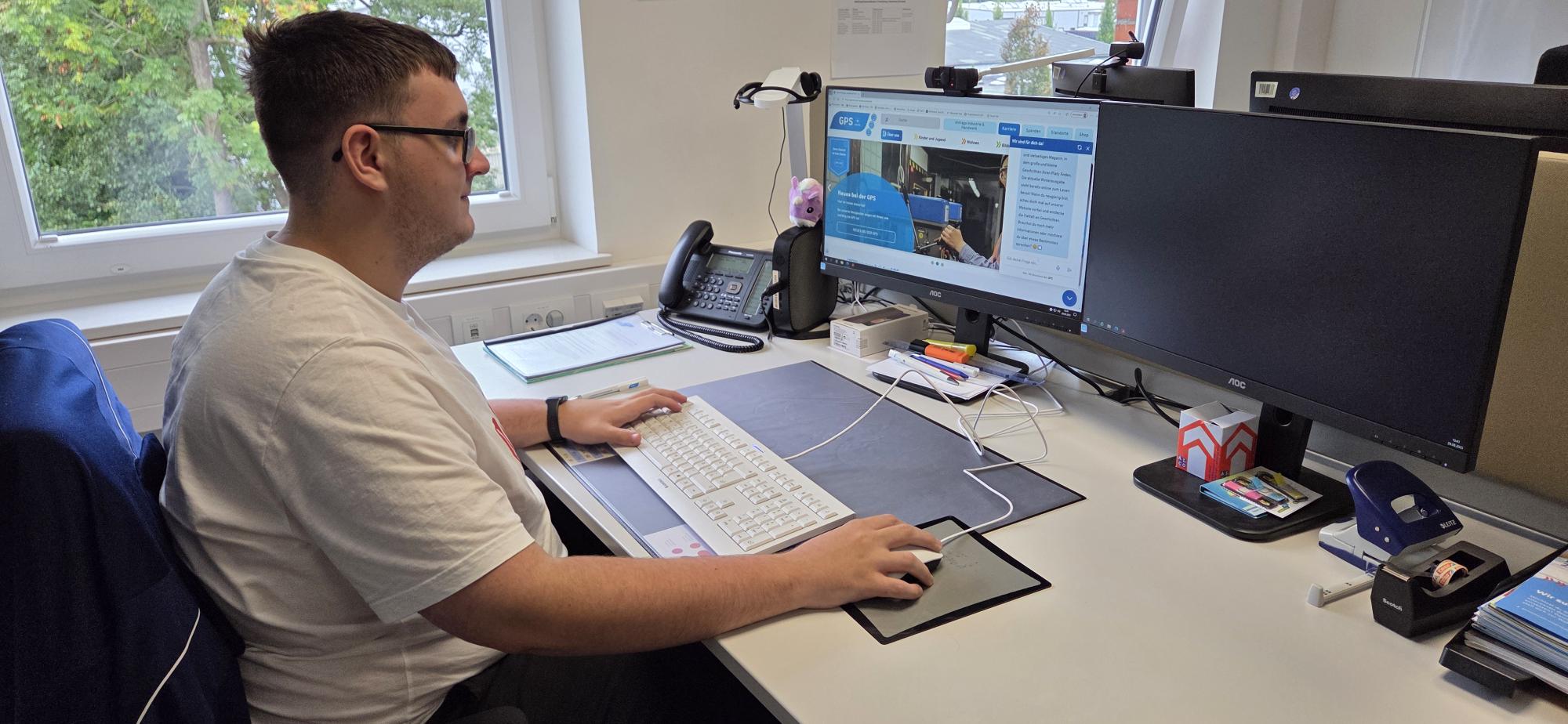 Ein junger Praktikant in der Abteilung "Kommunikation & Marketing" bei der GPS. Er sitzt an einem Schreibtisch und macht seine Aufgaben an einem Computer.
