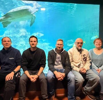 Gruppenfoto von fünf Menschen in einem Aquarium. Im Hintergrund sieht man eine große Schildkröte im Becken schwimmen.