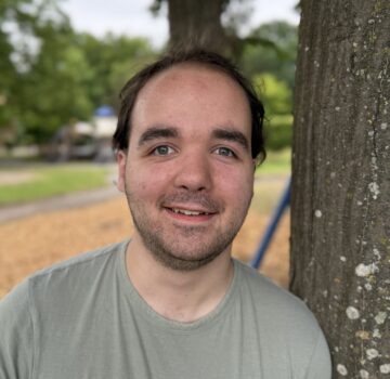 Ein junger Mann namens Sven Buhrs. Er hat dunkle kurze Haare und trägt ein graues T-Shirt und steht draußen vor der Kamera.