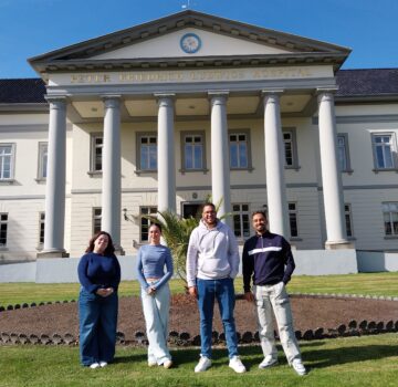 Vier junge Menschen draußen vor einem großen Gebäude. Die Sonne scheint.