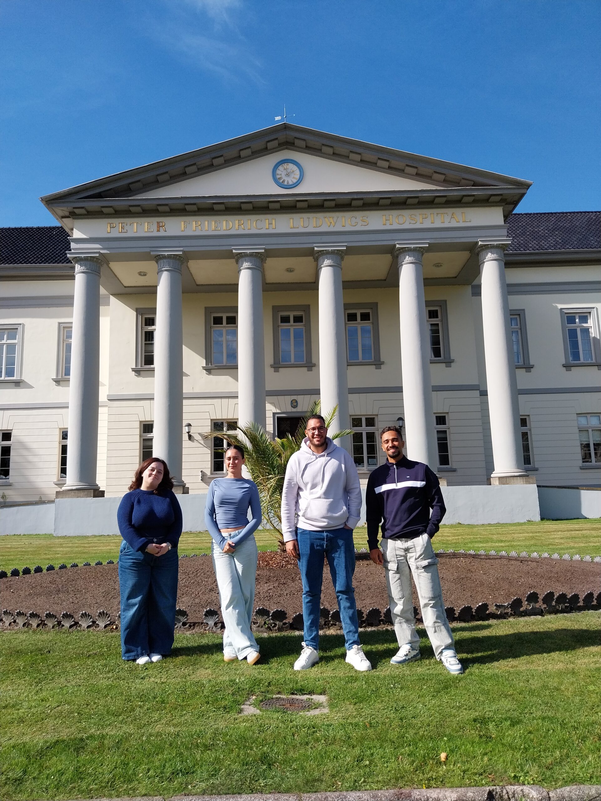 Vier junge Menschen draußen vor einem großen Gebäude. Die Sonne scheint.