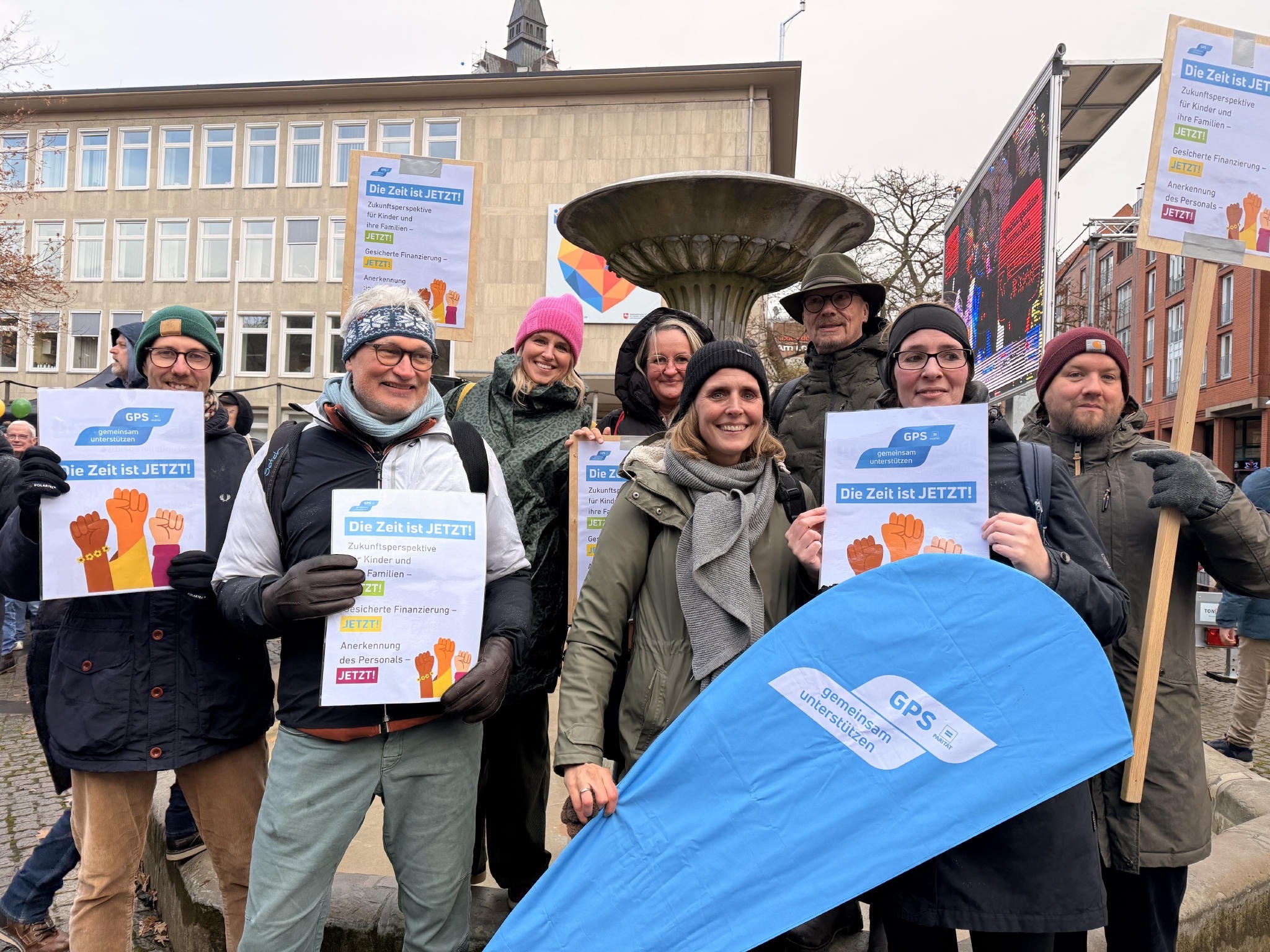 GPS Mitarbeiter und Mitarbeiterinnen bei einer Demonstration in einer Stadt draußen. Sie halten Plakate in der Hand.