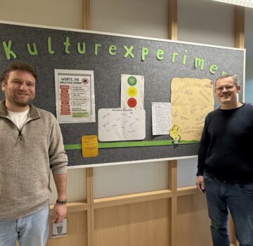 Zwei GPS Mitarbeiter stehen vor einer Tafel mit dem Thema "Kulturexperiment" in einem großen Raum.