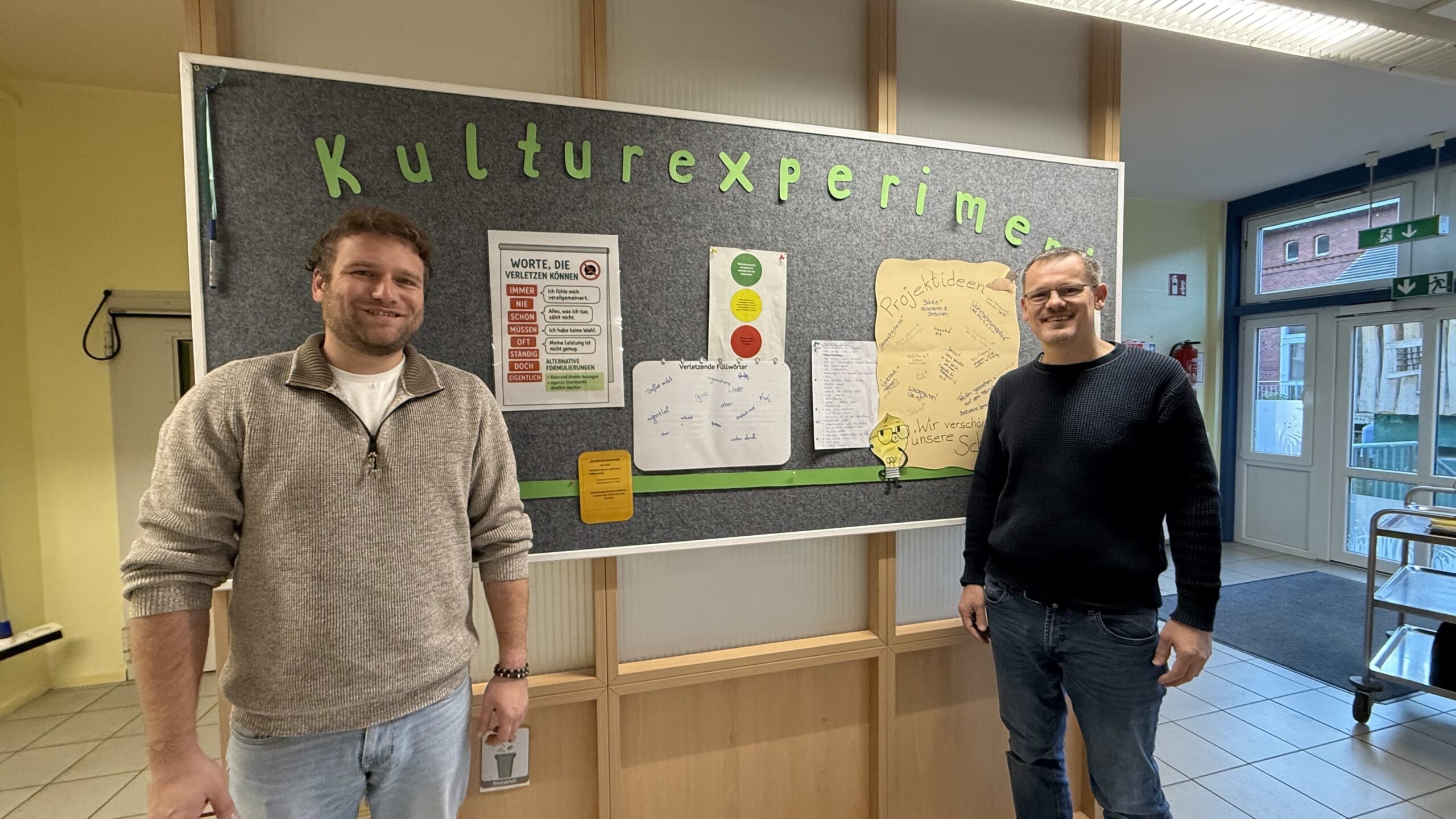 Zwei GPS Mitarbeiter stehen vor einer Tafel mit dem Thema "Kulturexperiment" in einem großen Raum.