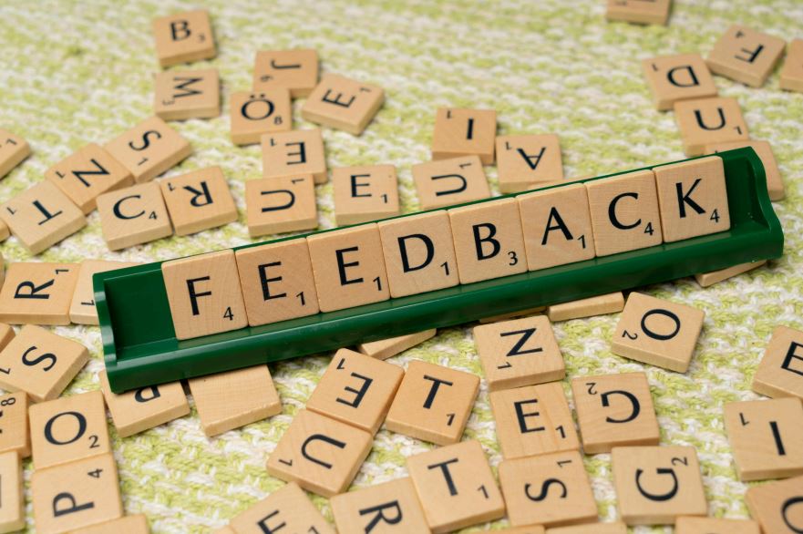Auf dem Bild sieht man ein grünes Schild und Holzschilder mit verschiedenen Buchstaben. Aus den Buchstaben liest man das Wort "Feedback"