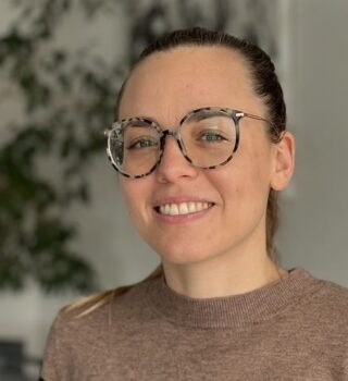 Eine Frau namens Theresa Orzessek mit dunklen langen Haaren mit Zopf und eine Brille. Sie trägt einen braun-schwarzen Pullover.