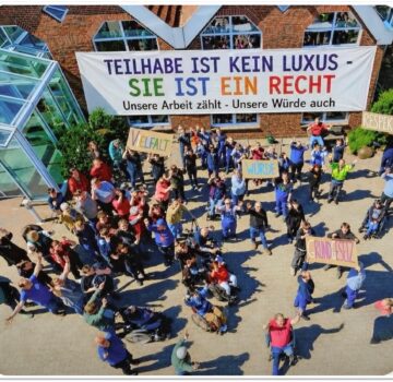 Viele Menschen von einer GPS Werkstatt draußen auf einem Gelände. Man sieht ein sehr langes großes Banner mit Bunt-Geschriebenes "Teilhabe ist kein Luxus - Sie ist ein Recht - Unsere Arbeit zählt - Unsere Würde auch".