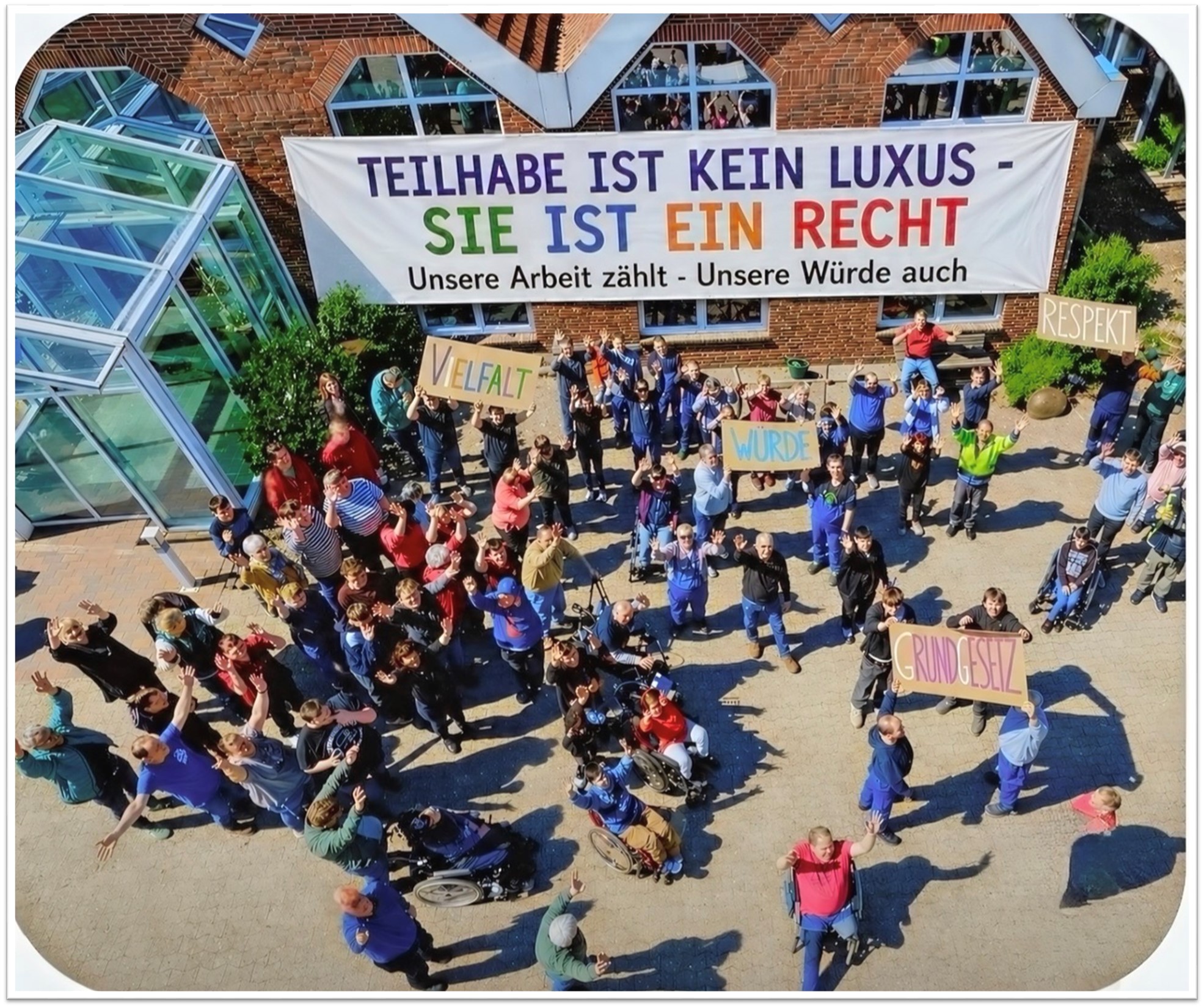 Viele Menschen von einer GPS Werkstatt draußen auf einem Gelände. Man sieht ein sehr langes großes Banner mit Bunt-Geschriebenes "Teilhabe ist kein Luxus - Sie ist ein Recht - Unsere Arbeit zählt - Unsere Würde auch".