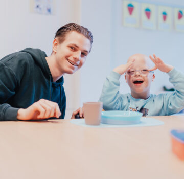 Ein junger Mann und ein kleiner Junge mit einer Behinderung sitzen an einem Tisch in einem Raum.