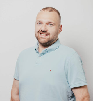 Ein Mann namens Christian Alberts mit kurzen dunklen Haaren und einen Vollbart. Sie trägt einen hell-blauen Pullover.