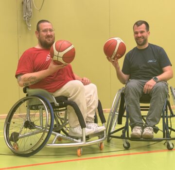 Zwei Männer in einem Rollstuhl in einer Sporthalle. Beide halten einen Basketball in ihrer Hand.