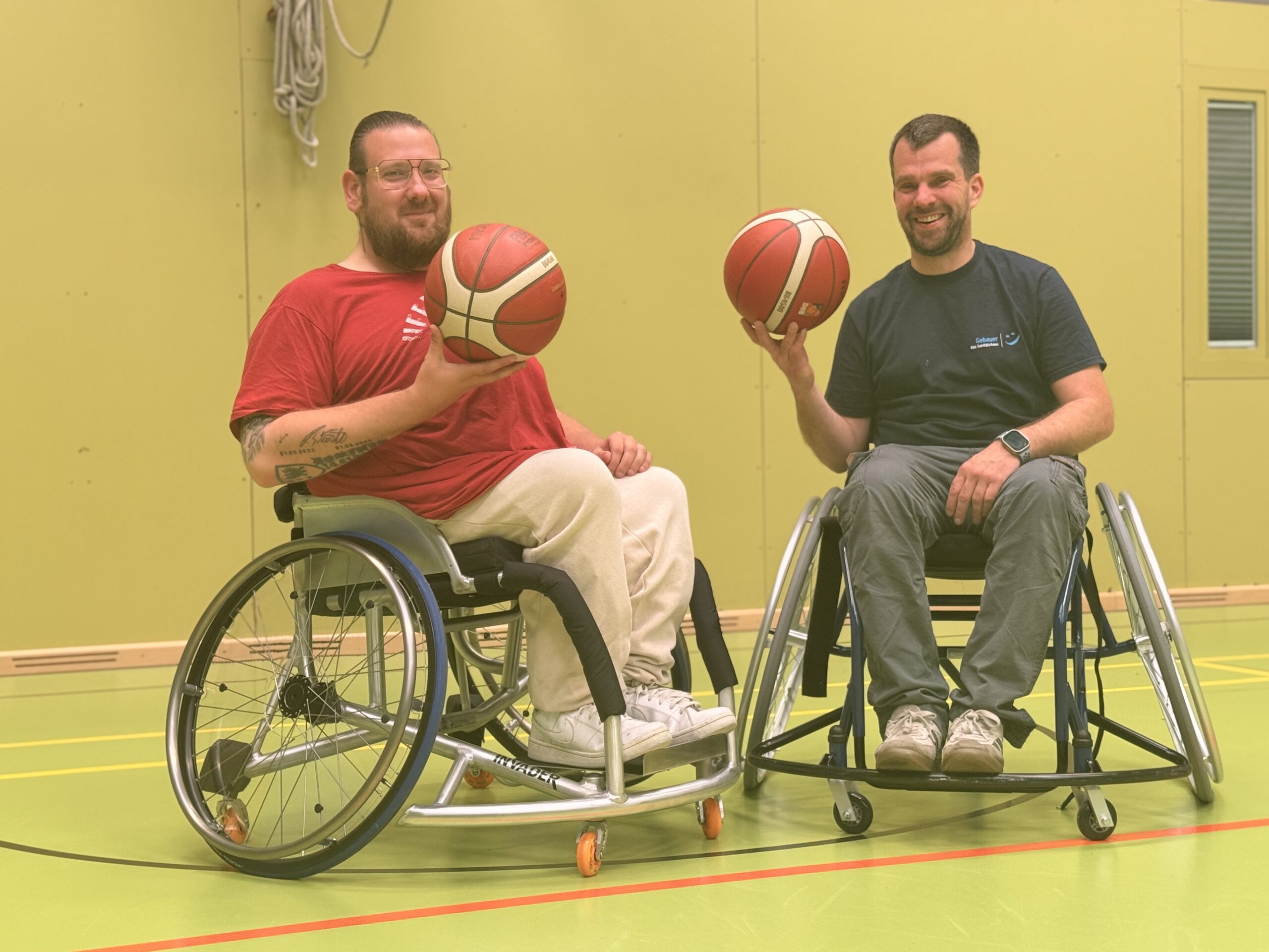 Zwei Männer in einem Rollstuhl in einer Sporthalle. Beide halten einen Basketball in ihrer Hand.
